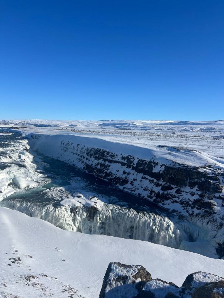 Vista panorámica de una gran cascada parcialmente congelada rodeada de nieve y acantilados bajo un cielo azul claro.