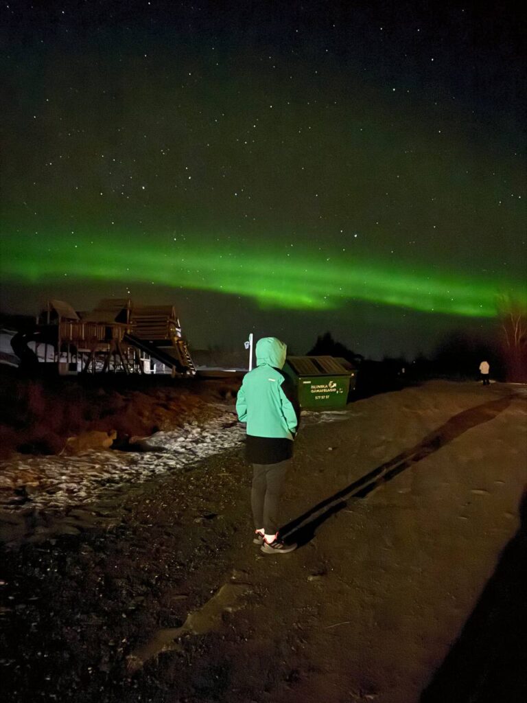 Persona observando una aurora boreal verde en el cielo nocturno, con estrellas visibles y construcciones al fondo.