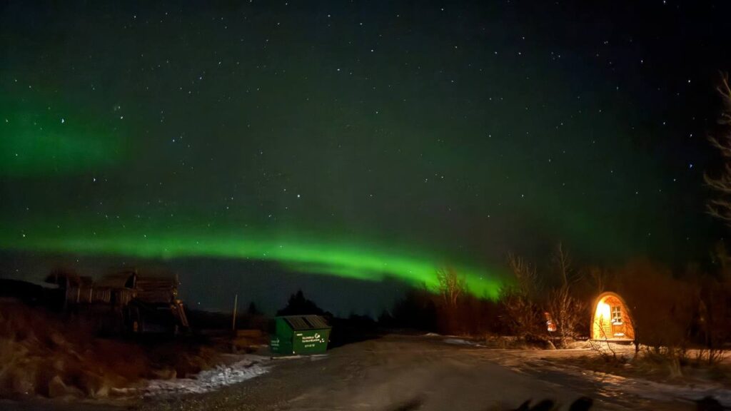 Auroras boreales verdes iluminan el cielo nocturno estrellado sobre un camino rural con una cabaña iluminada y árboles al fondo.