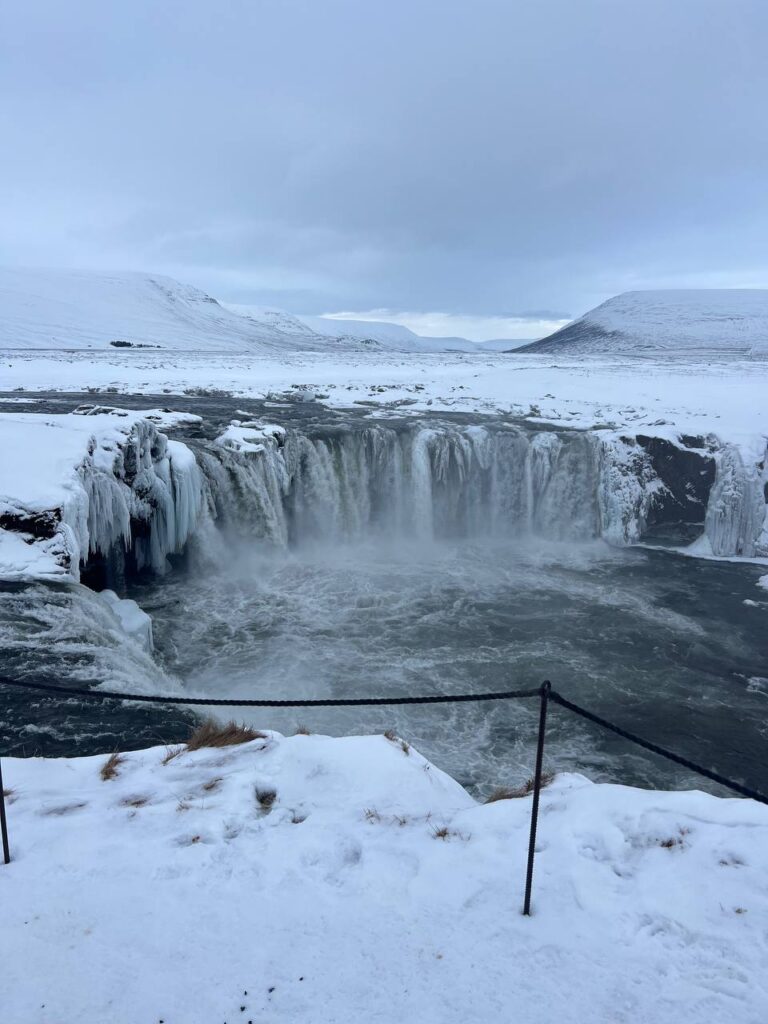 Vista frontal de una cascada parcialmente congelada rodeada de nieve en un paisaje montañoso invernal.