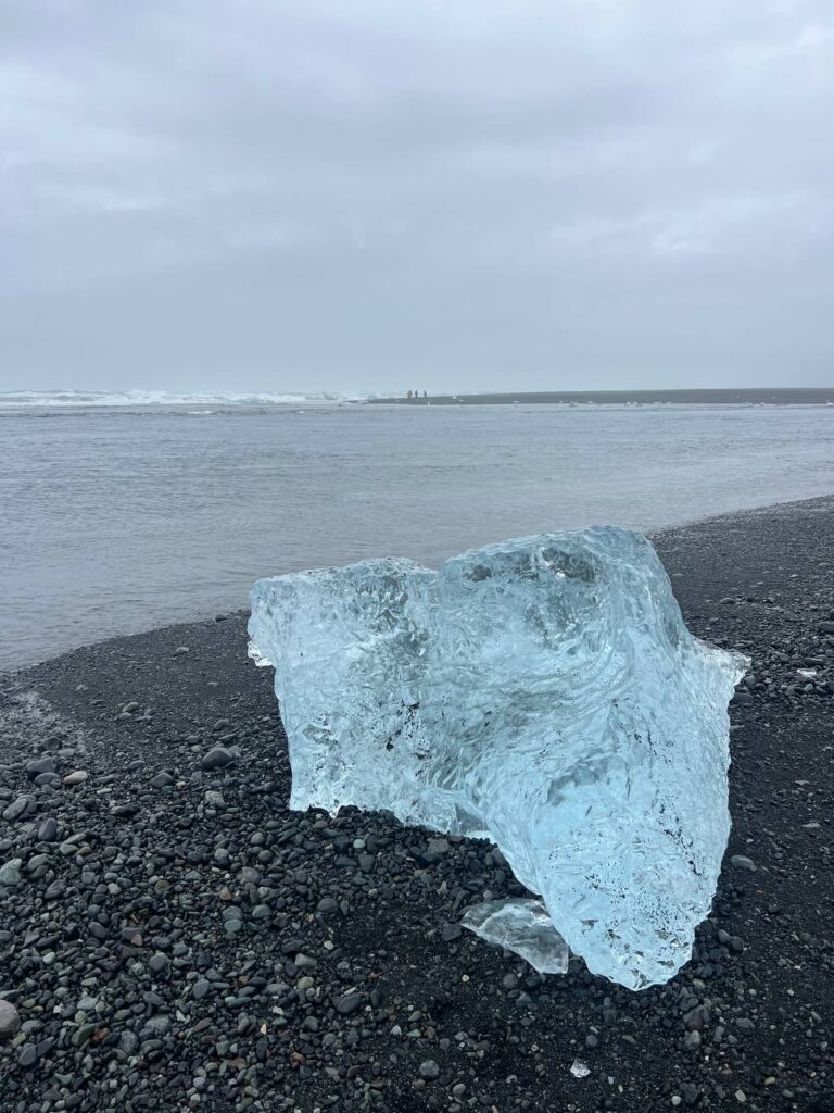 Gran bloque de hielo translúcido sobre la arena volcánica negra de una playa junto al océano.