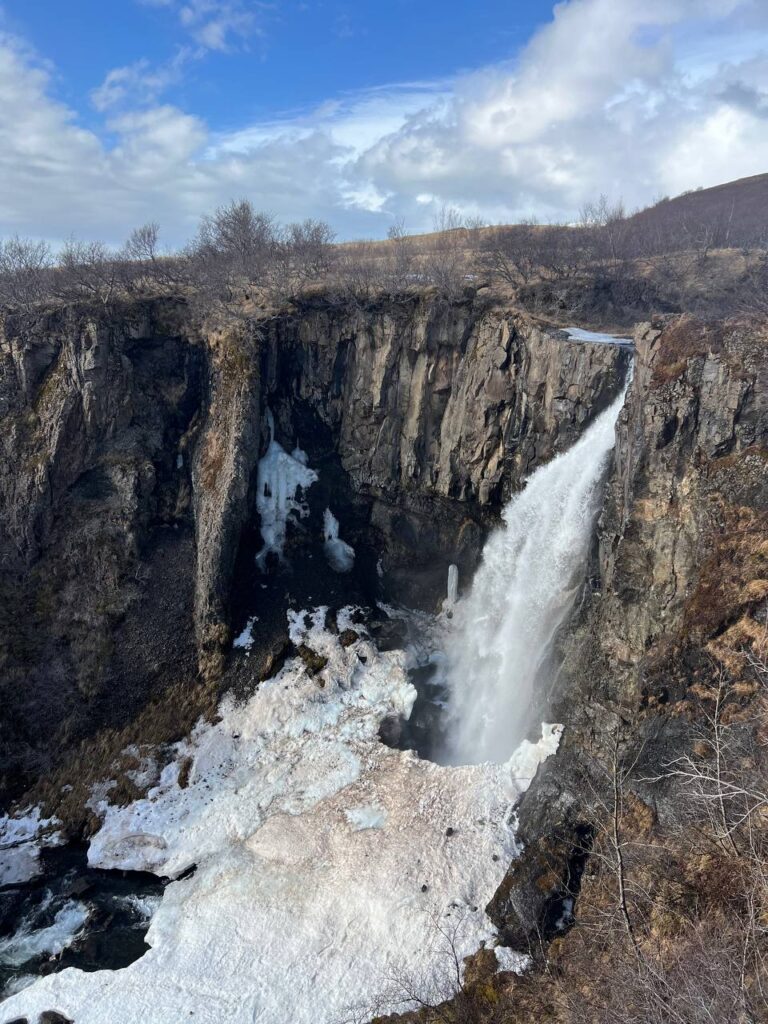 Cascada cayendo por un acantilado rocoso con restos de hielo y nieve al pie, rodeada de árboles sin hojas y cielo parcialmente nublado.
