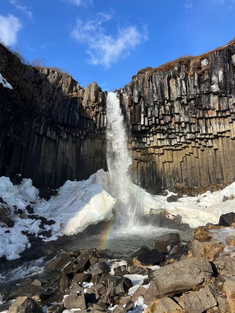 Cascada descendiendo entre columnas de basalto con hielo en la base y un pequeño arcoíris en el agua.