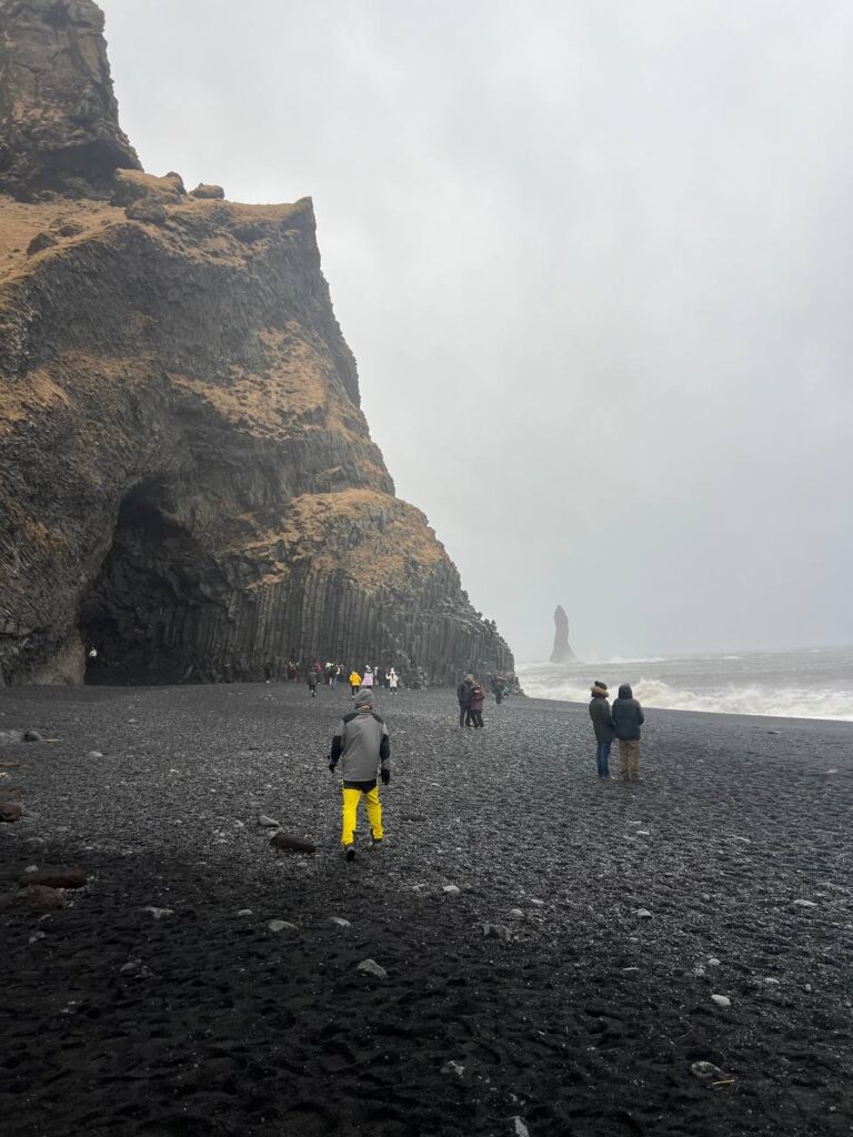 Personas caminando por una playa de arena negra con formaciones basálticas y un mar embravecido al fondo, en un día nublado.