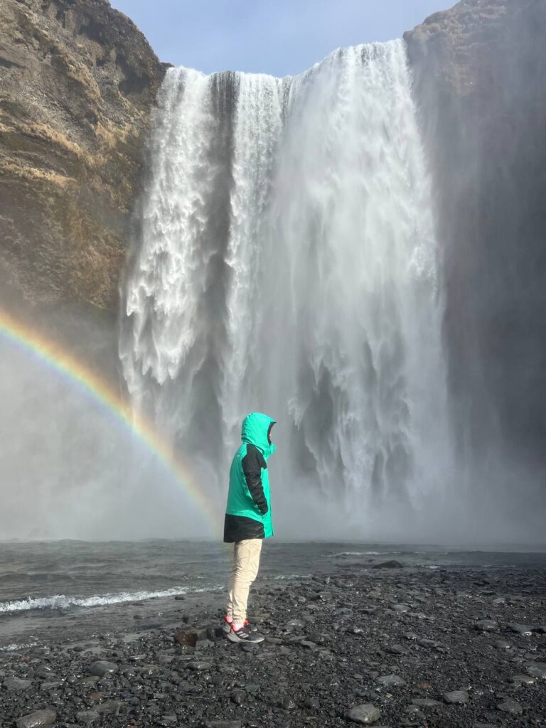 Persona con chaqueta verde observando una gran cascada con un arcoíris a su lado, en un paisaje de rocas oscuras.