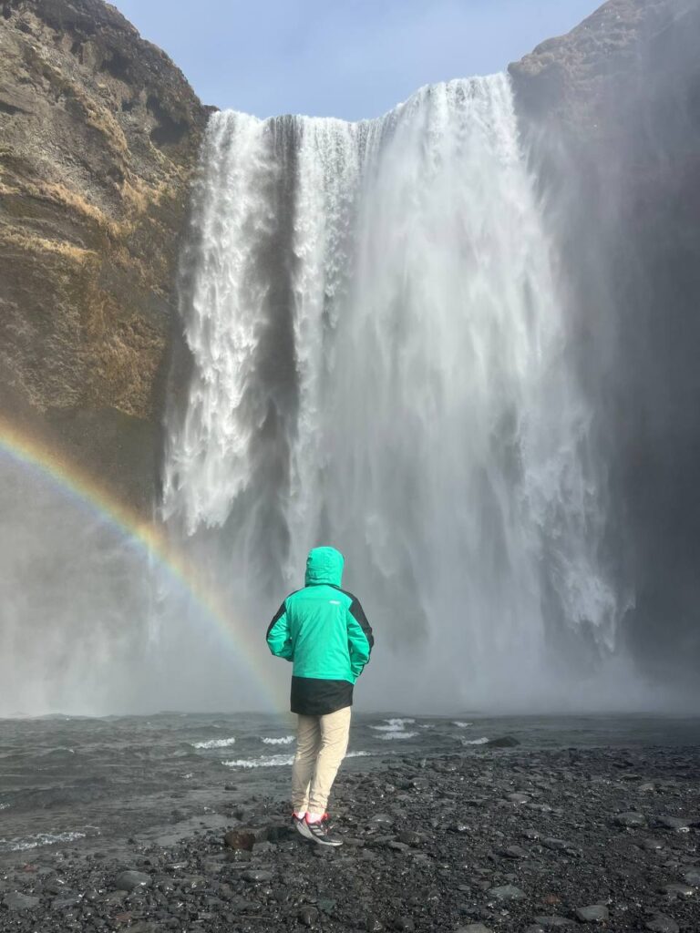 Persona de espaldas observando una gran cascada con un arcoíris formado por la bruma del agua, en un entorno rocoso.