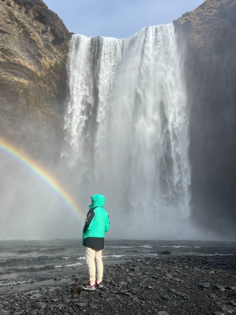 Persona con chaqueta verde observando una cascada imponente con un arcoíris formado por la niebla del agua.