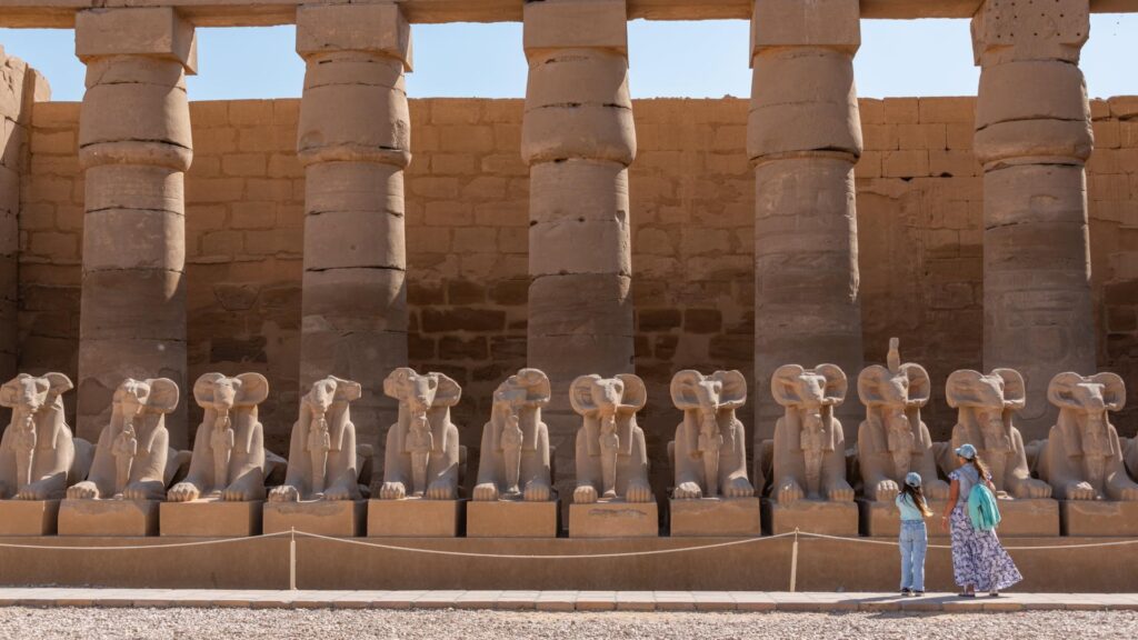 Esculturas de carneros en fila frente a columnas egipcias en el Templo de Karnak, con dos turistas observándolas.