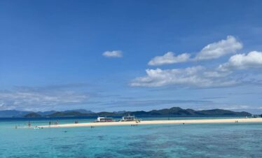 Isla de arena blanca rodeada por aguas turquesas y transparentes, con personas disfrutando del mar y embarcaciones ancladas al fondo.