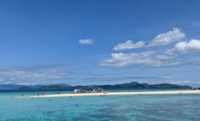 Isla de arena blanca rodeada por aguas turquesas y transparentes, con personas disfrutando del mar y embarcaciones ancladas al fondo.