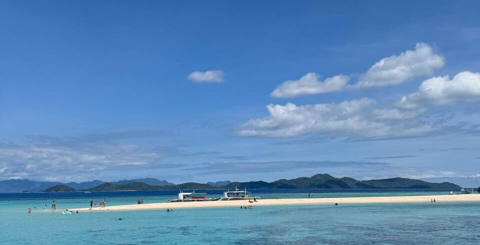 Isla de arena blanca rodeada por aguas turquesas y transparentes, con personas disfrutando del mar y embarcaciones ancladas al fondo.