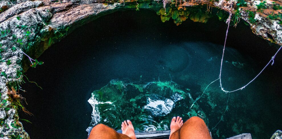 Persona sentada al borde de un cenote natural con los pies colgando sobre el agua cristalina rodeada de rocas y vegetación.