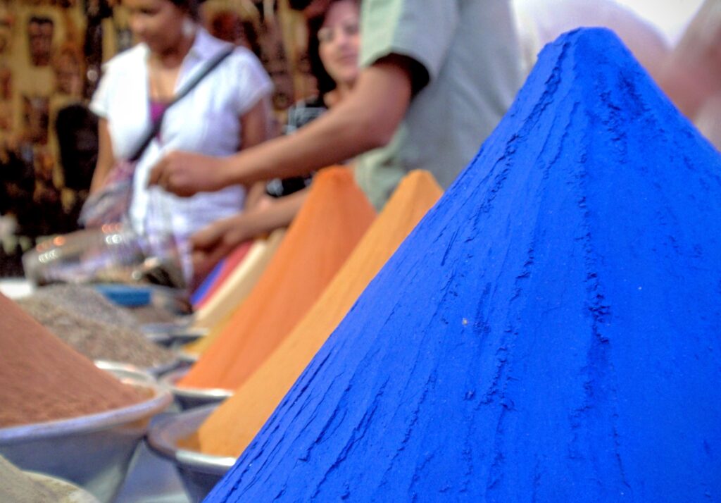 Conos de pigmentos de colores intensos, como azul y naranja, en un mercado con personas de fondo.