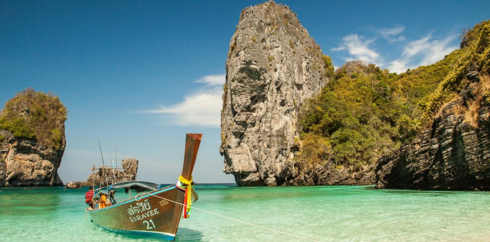Barco tradicional tailandés anclado en aguas turquesas junto a formaciones rocosas y acantilados en una playa tropical.