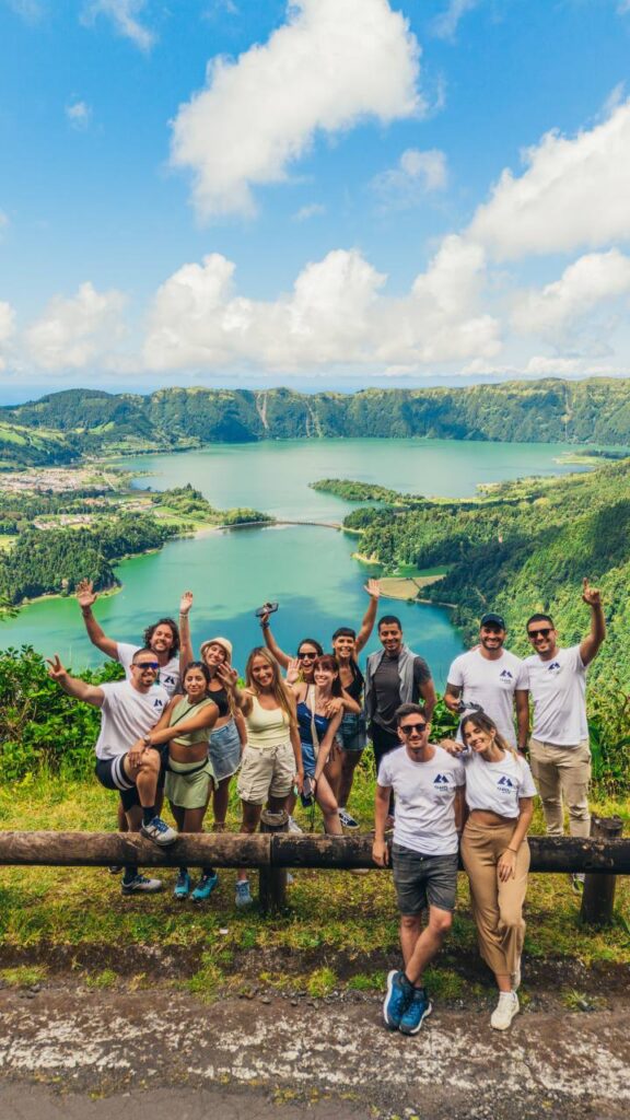 Grupo de personas sonrientes posando frente a la Laguna de las Siete Ciudades en las Azores, Portugal, con un paisaje verde y montañoso al fondo.