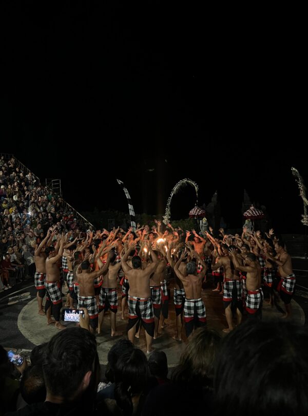 Grupo de hombres con sarong a cuadros negros y rojos realizando una danza tradicional balinesa en círculo alrededor del fuego, con espectadores observando de noche.