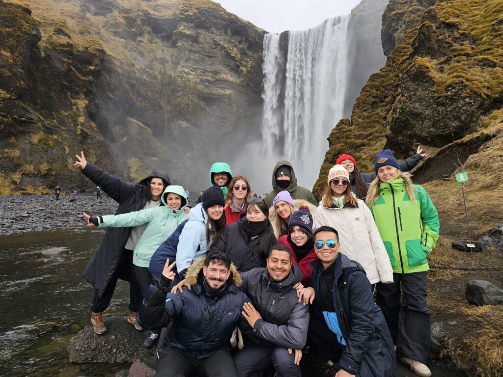 Grupo de personas posando frente a una gran cascada rodeada de montañas cubiertas de musgo.