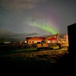 Aurora boreal iluminando el cielo nocturno sobre una casa de campo y una excavadora en primer plano.