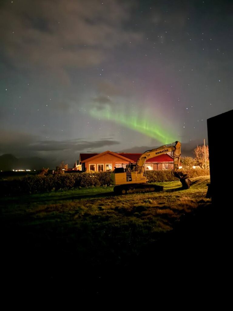 Aurora boreal iluminando el cielo nocturno sobre una casa de campo y una excavadora en primer plano.