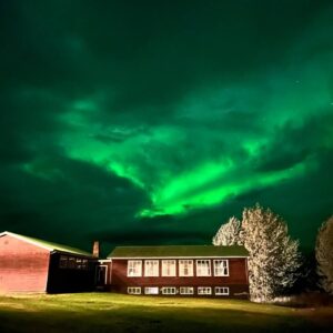 Casa de madera iluminada bajo un cielo nocturno con aurora boreal verde brillante.