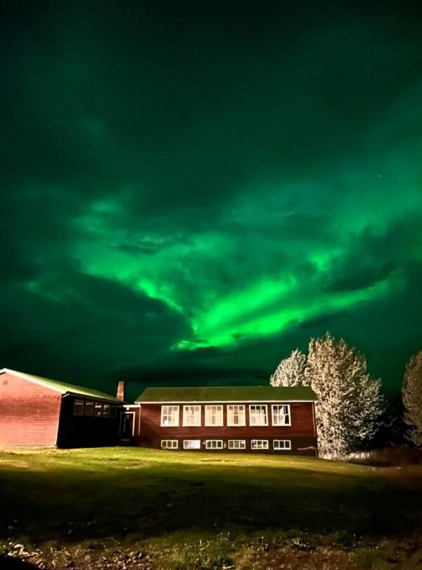 Casa de madera iluminada bajo un cielo nocturno con aurora boreal verde brillante.