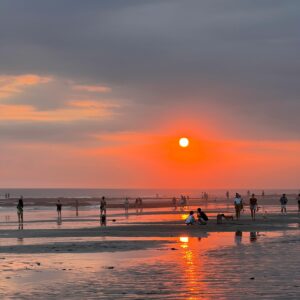 Personas disfrutando de un atardecer en la playa con el sol reflejado en la arena mojada.