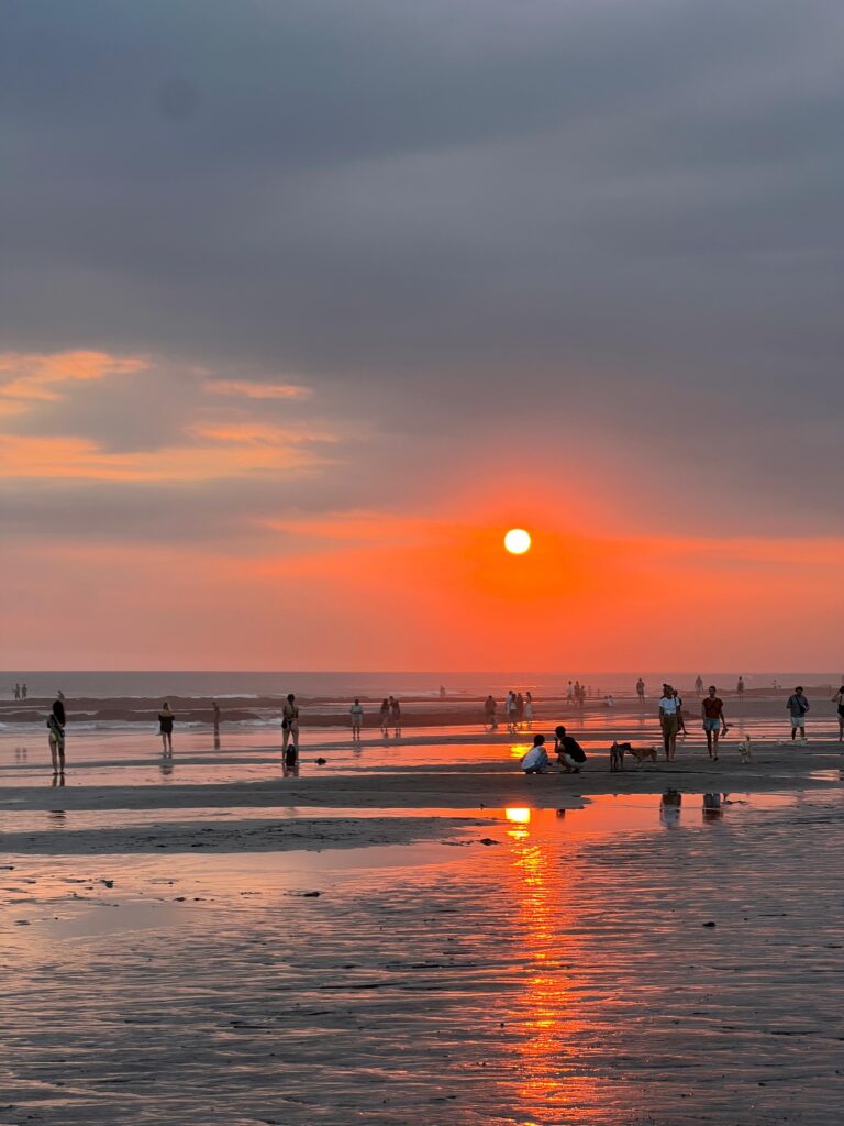 Personas disfrutando de un atardecer en la playa con el sol reflejado en la arena mojada.