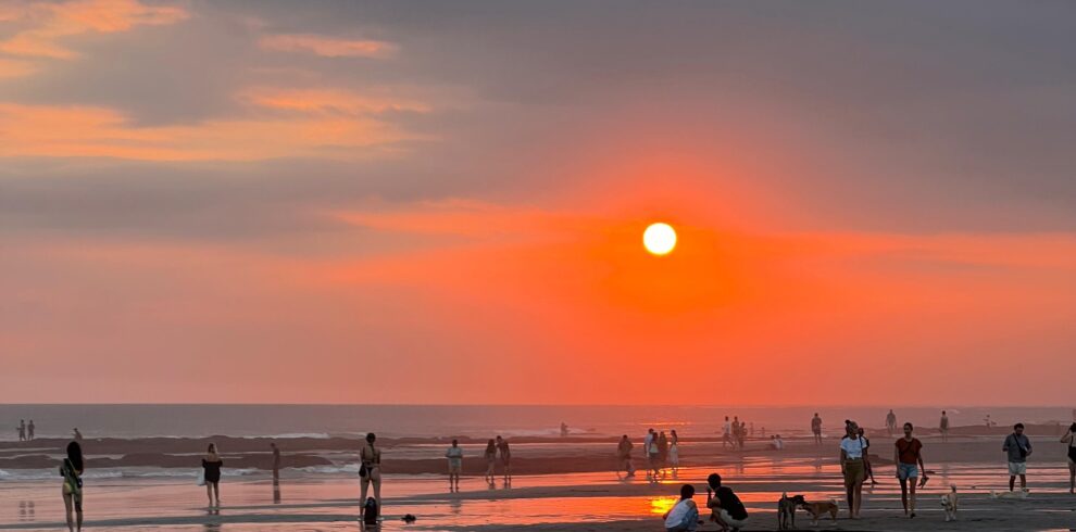 Personas disfrutando de un atardecer en la playa con el sol reflejado en la arena mojada.