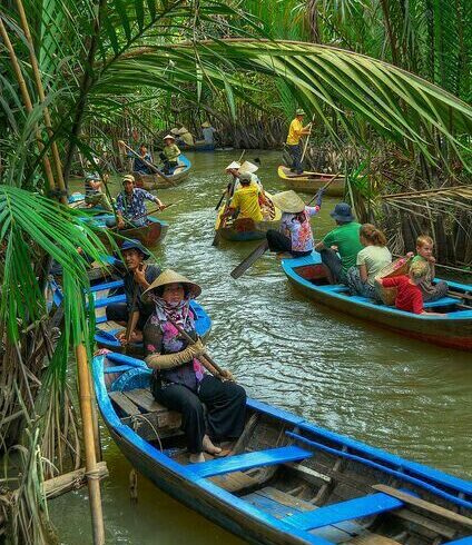 Personas navegando en pequeñas barcas de remo a través de un canal rodeado de vegetación tropical densa, posiblemente en el delta del Mekong, Vietnam.