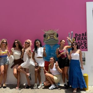 Grupo de diez mujeres posando sonrientes frente a un mural color rosa con un dibujo de un pez con camisa azul y la frase "Debí tirar más fotos".