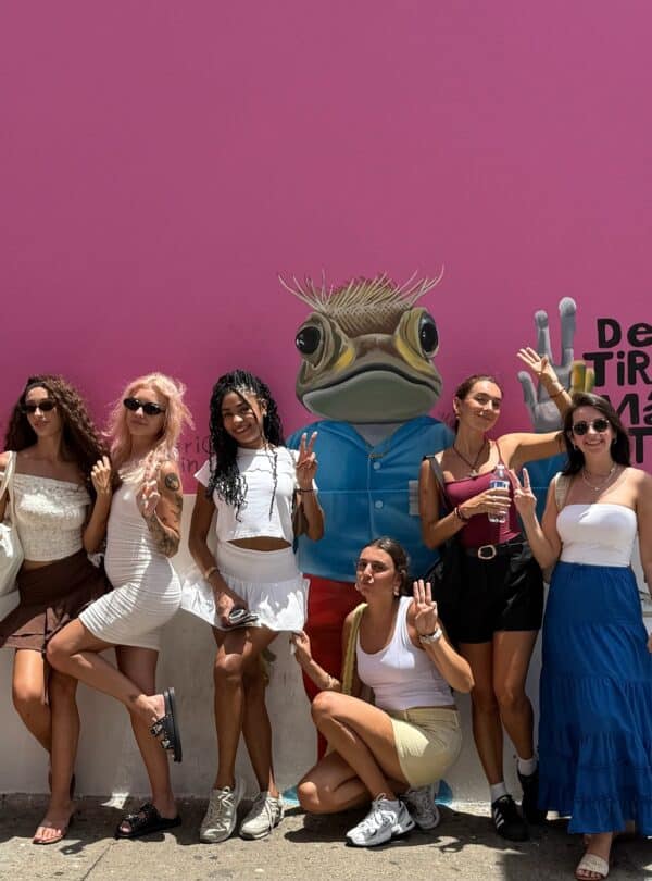 Grupo de diez mujeres posando sonrientes frente a un mural color rosa con un dibujo de un pez con camisa azul y la frase "Debí tirar más fotos".