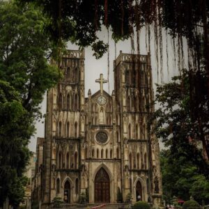 Fachada gótica de la Catedral de San José en Hanói, Vietnam, con árboles frondosos alrededor y vehículos circulando frente al edificio.