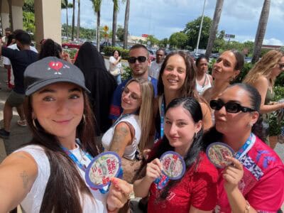 Grupo de mujeres sonrientes posando con boletos o souvenirs del "Bad Bunny World’s Hottest Tour" mientras hacen fila en un espacio al aire libre.