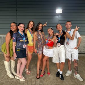 Grupo de jóvenes posando con atuendos coloridos y credenciales colgadas al cuello, frente a un muro gris metálico en lo que parece ser la entrada a un evento.
