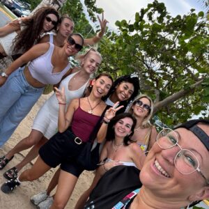Grupo de mujeres sonrientes posando para una selfie en un entorno tropical con árboles y arena, luciendo ropa veraniega.