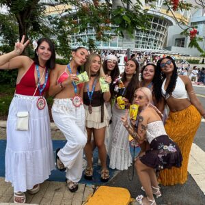 Grupo de mujeres jóvenes posando con vestimenta veraniega y colorida frente a un centro de eventos, sosteniendo snacks y bebidas.