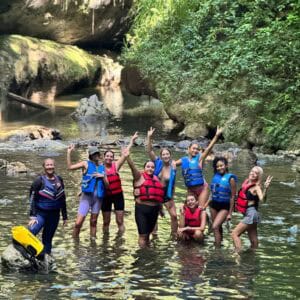 Grupo de mujeres usando chalecos salvavidas posando felices dentro de un río rodeado de vegetación y formaciones rocosas en un entorno selvático.