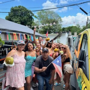 Grupo de personas posando felices frente a un puesto callejero con cocos y una furgoneta, ondeando una bandera de Puerto Rico bajo un cielo azul.