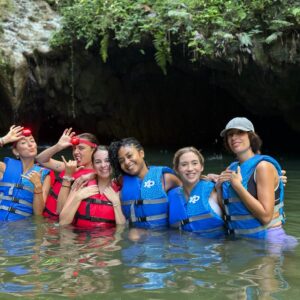 Grupo de seis mujeres con chalecos salvavidas posando felices en el agua frente a la entrada de una cueva natural rodeada de vegetación.