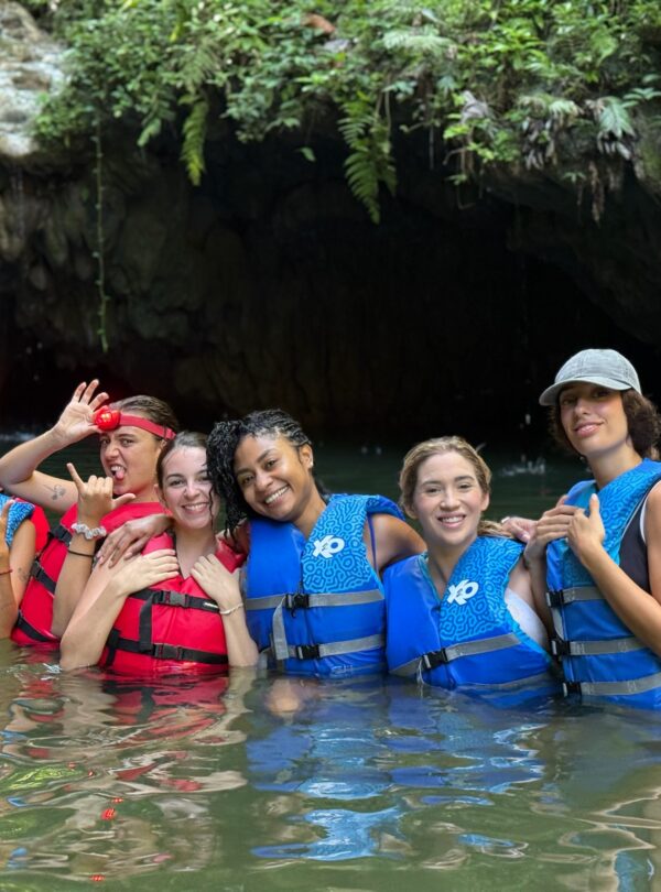 Grupo de seis mujeres con chalecos salvavidas posando felices en el agua frente a la entrada de una cueva natural rodeada de vegetación.