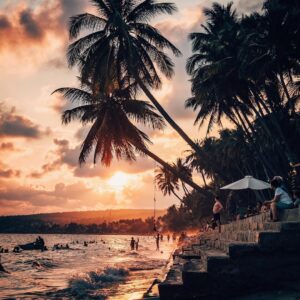 Playa tropical al atardecer con palmeras, bañistas en el mar y personas sentadas en escalones junto a la costa.