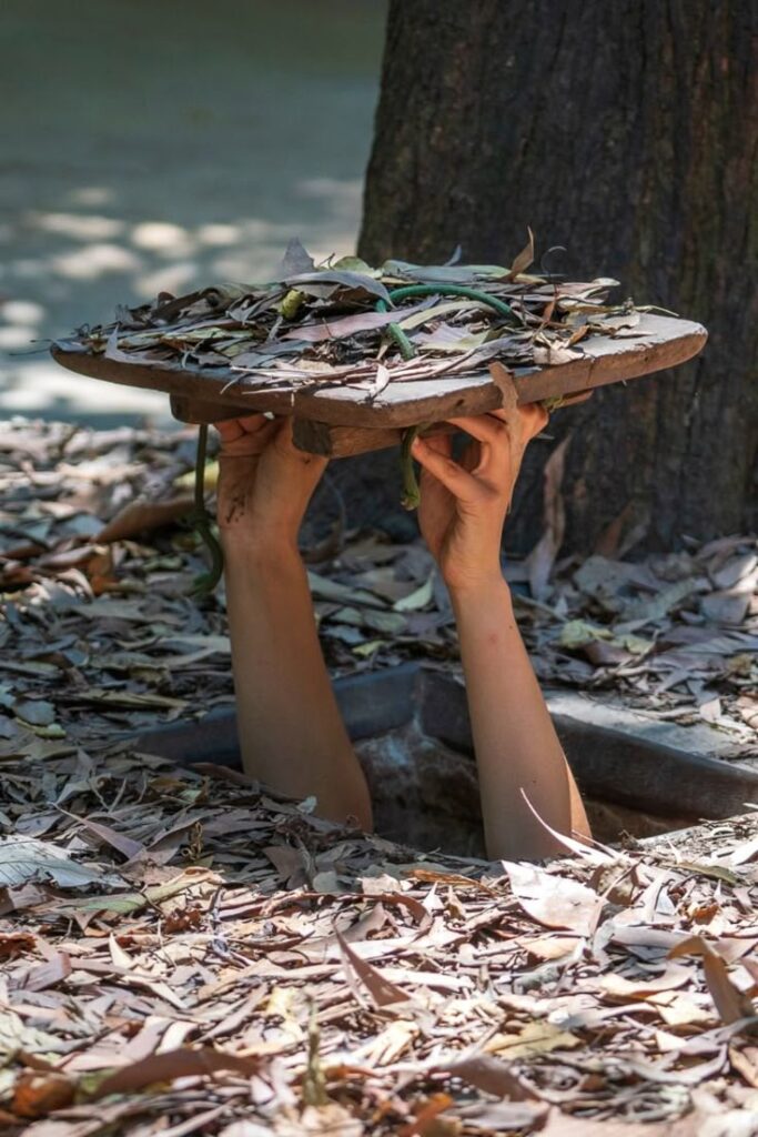 Manos emergen de un túnel subterráneo sosteniendo una tapa camuflada con hojas secas en la selva.