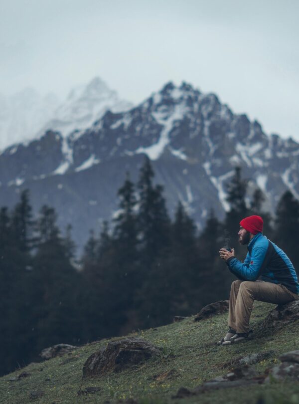 Hombre sentado al aire libre en una montaña, con gorro rojo y chaqueta azul, bebiendo de una taza caliente, con pinos y montañas nevadas al fondo.
