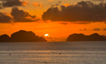 Atardecer naranja sobre el mar con el sol ocultándose entre dos islas y nubes oscuras en el cielo.