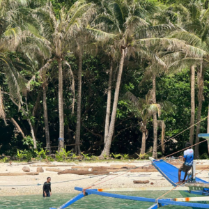 Hombre junto a un barco tradicional en aguas turquesas frente a una playa con palmeras y vegetación tropical.