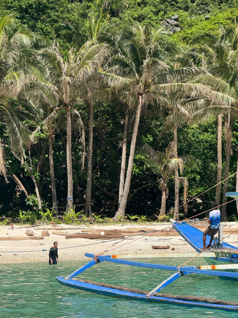 Hombre junto a un barco tradicional en aguas turquesas frente a una playa con palmeras y vegetación tropical.