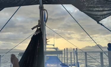 Vista desde una hamaca en la cubierta de un barco al atardecer, con el mar y montañas en el horizonte.