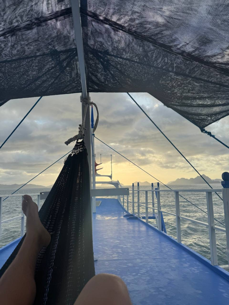 Vista desde una hamaca en la cubierta de un barco al atardecer, con el mar y montañas en el horizonte.