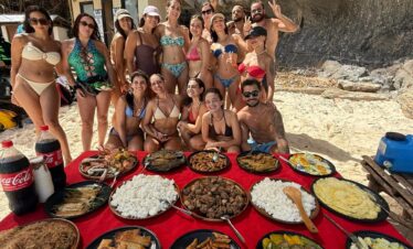Grupo de amigos en la playa posando detrás de una mesa con comida variada durante una comida al aire libre.