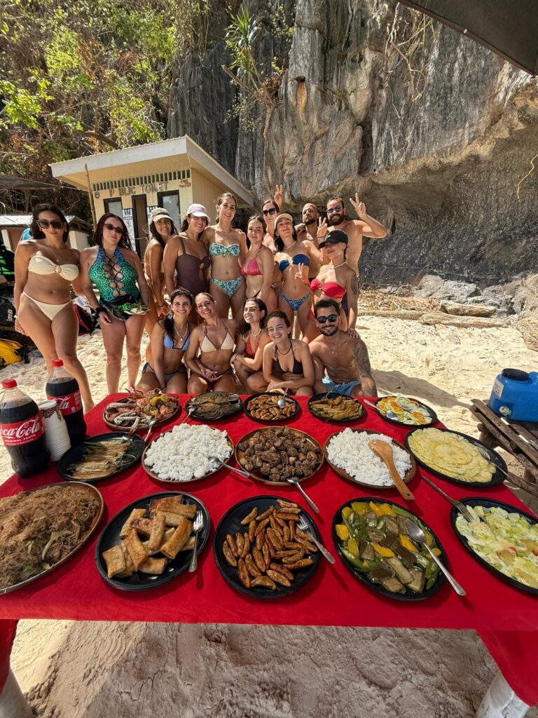 Grupo de amigos en la playa posando detrás de una mesa con comida variada durante una comida al aire libre.